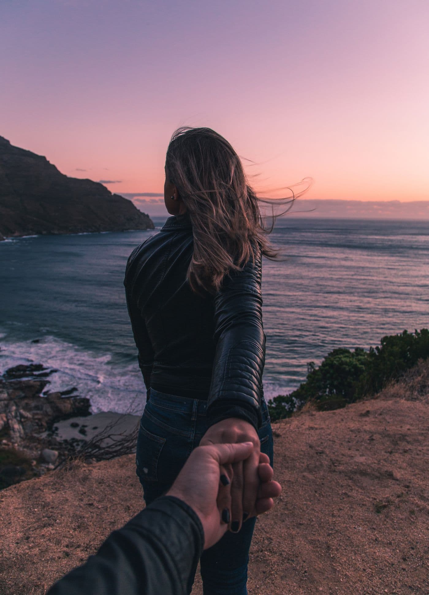 A woman leading someone by the hand toward a cliff overlooking the ocean at sunset