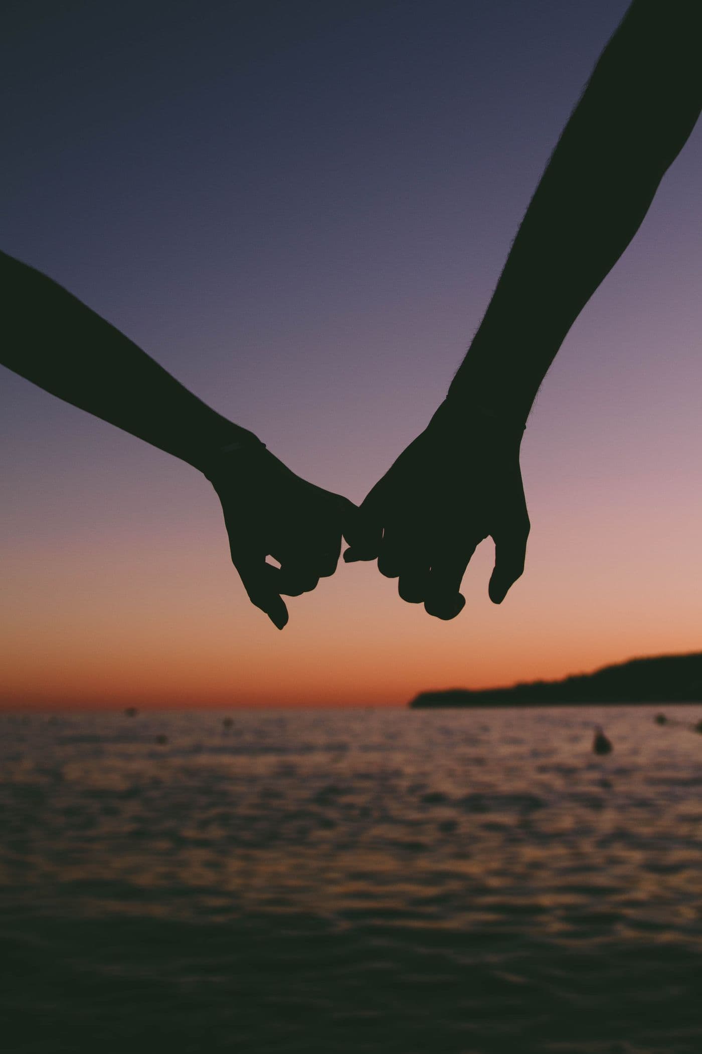 Two hands linked by the pinkies against a sunset over the water